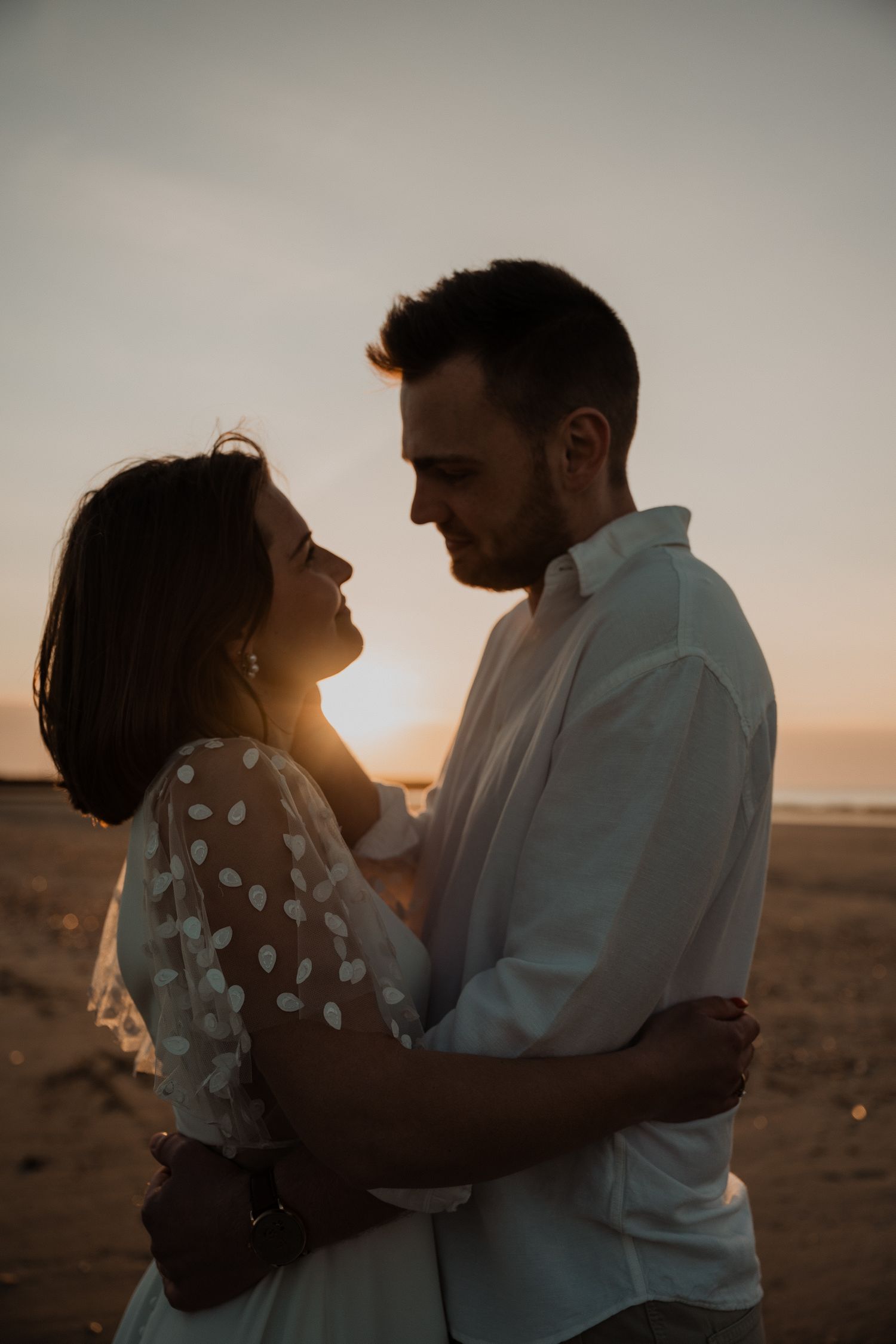 Couple embraces in romantic sunset moment on beach with polka dot dress and white shirt silhouetted against golden sky.