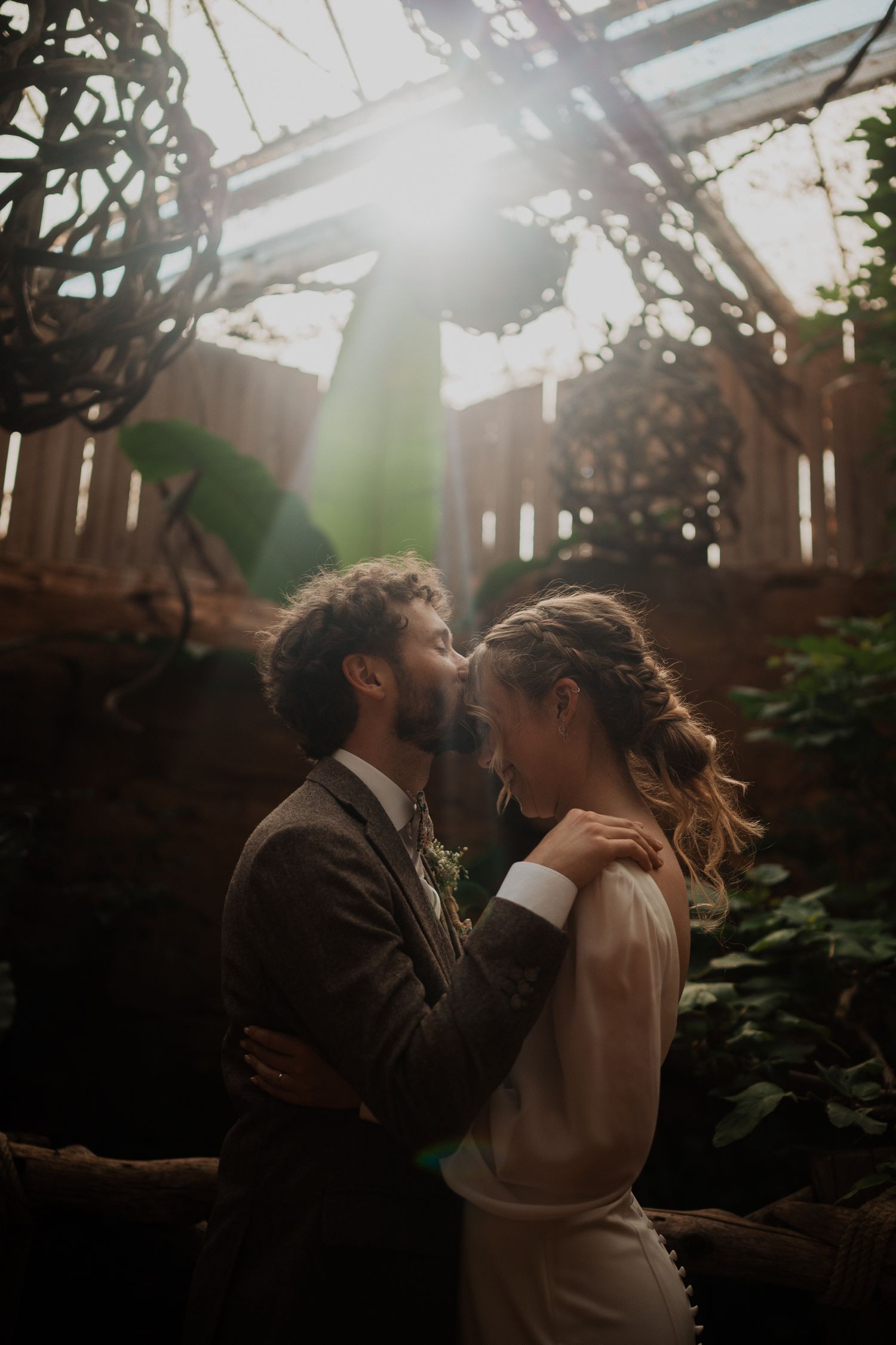 A romantic couple embraces in a garden backyard with sunlight streaming through a wooden fence creating a moody atmosphere.