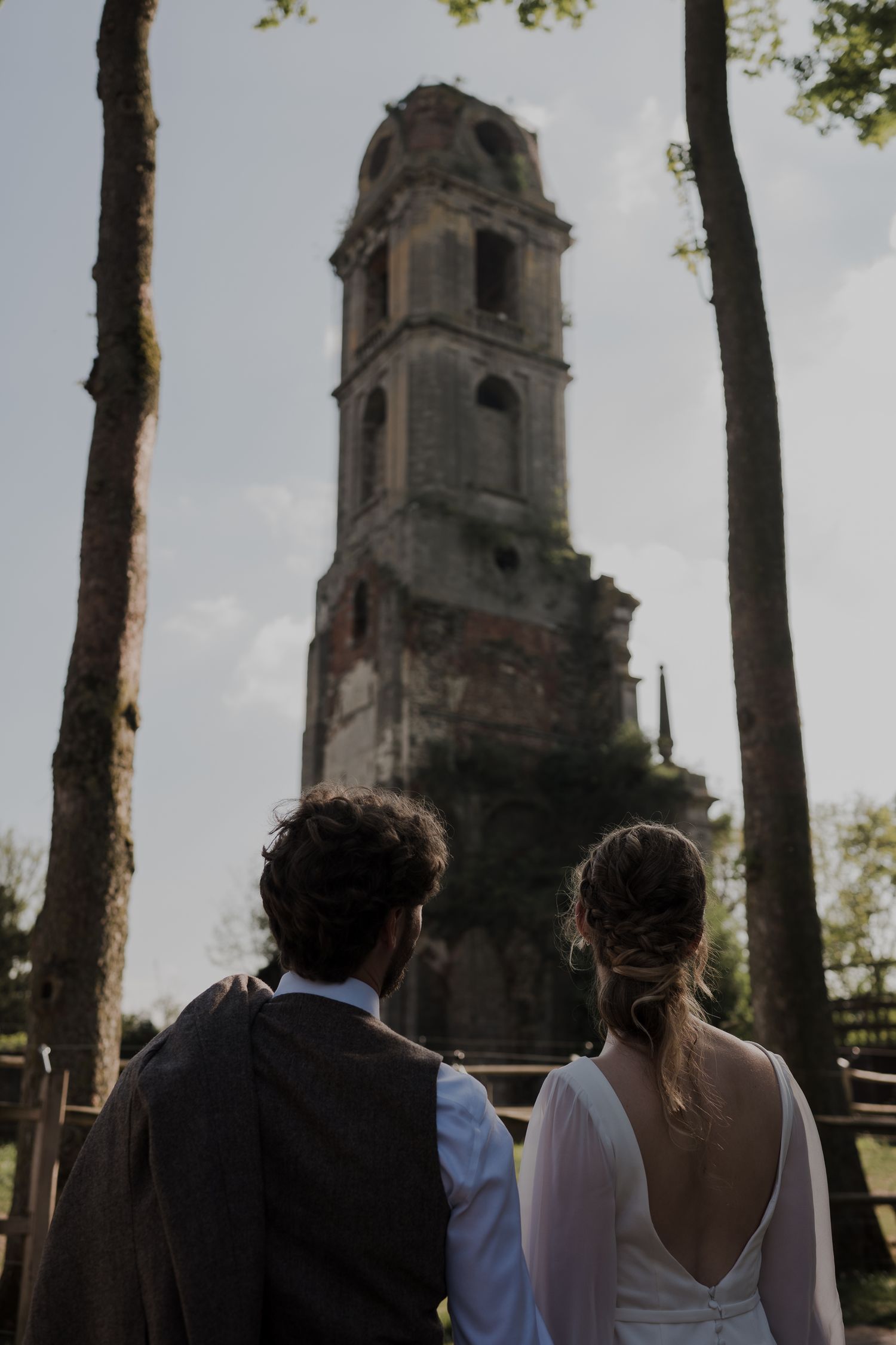 A couple stands together looking at a tall historic stone bell tower framed between trees.