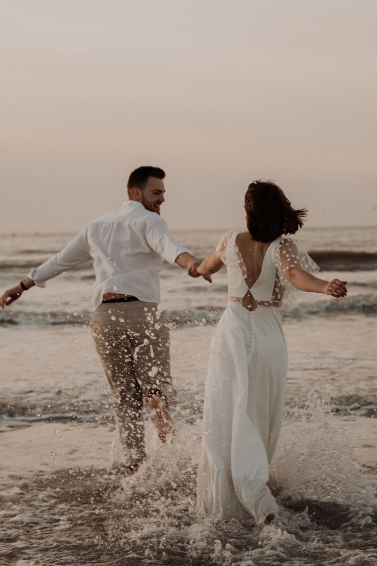Newlywed couple splashing and running through shallow ocean water at sunset in wedding attire.