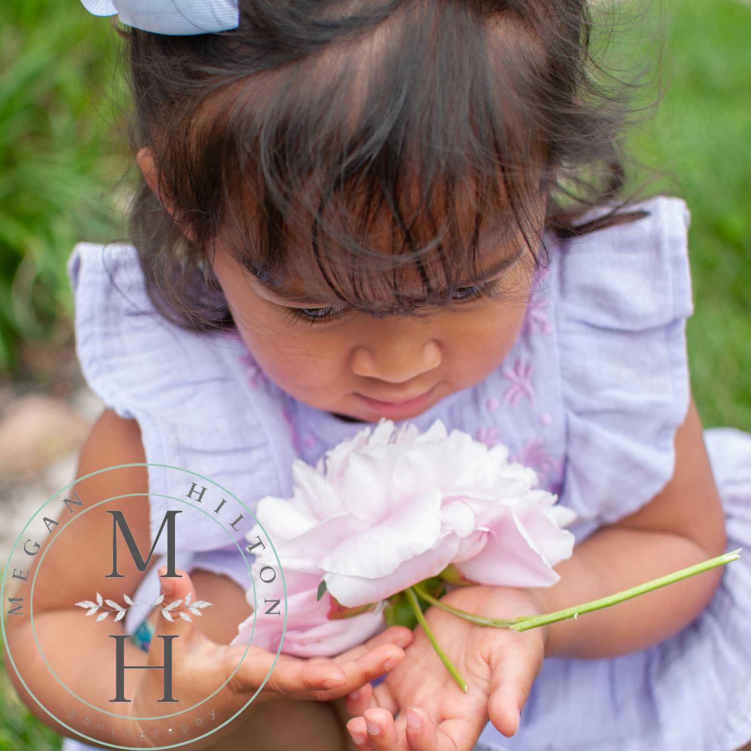 A small child in a lavender dress leaning down to smell a delicate pink peony flower in a garden setting.