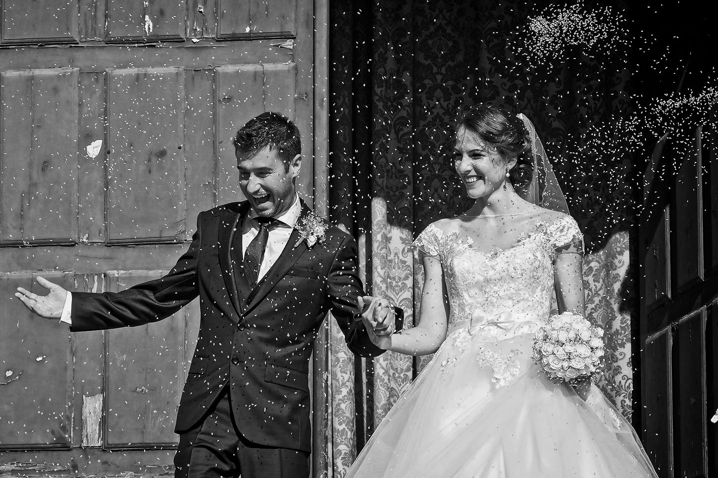 Black and white photo of newlyweds celebrating with confetti and joyful gestures outside a church building.
