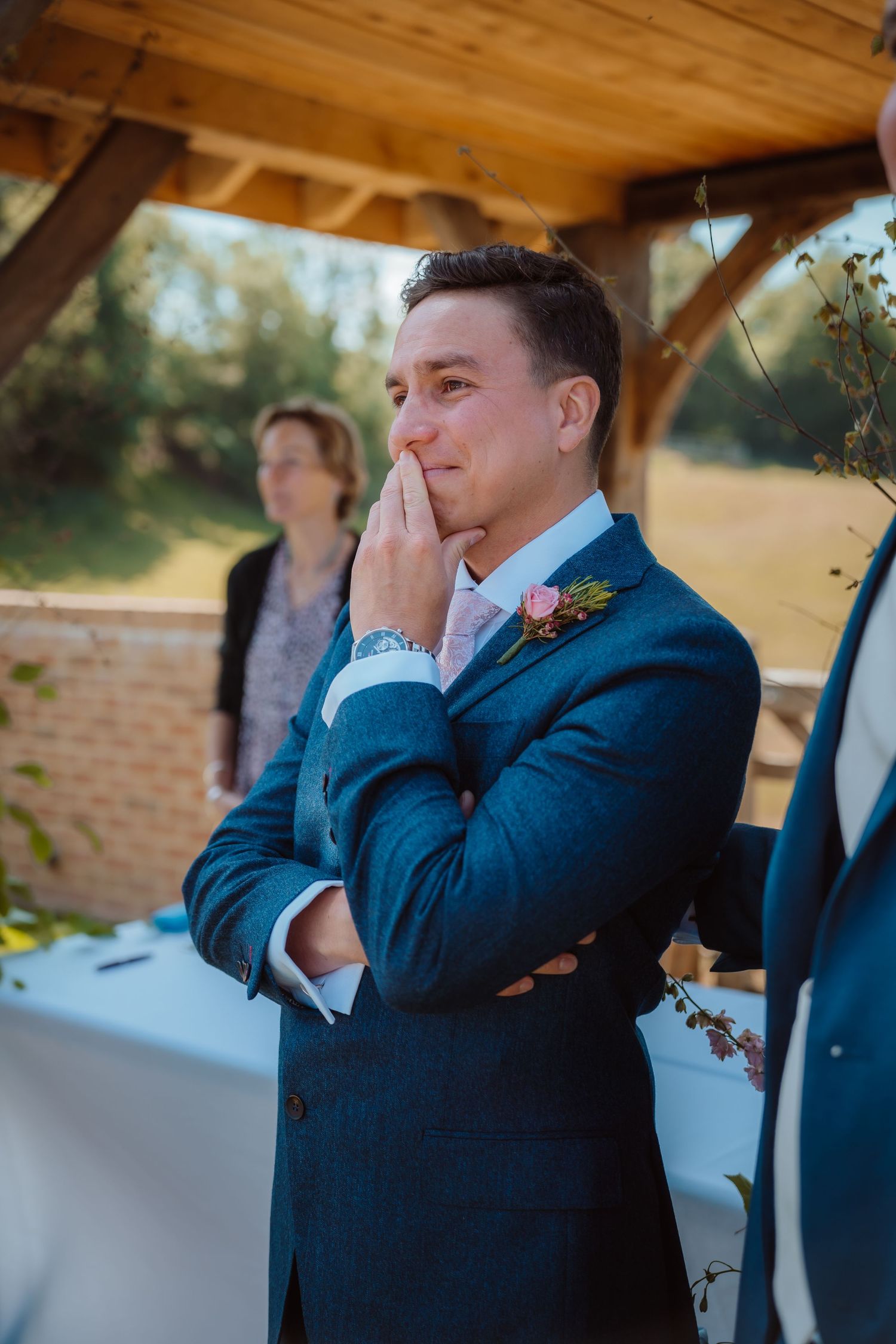 An emotional moment at a wedding ceremony as someone in a navy blue suit stands thoughtfully under a wooden arbor.