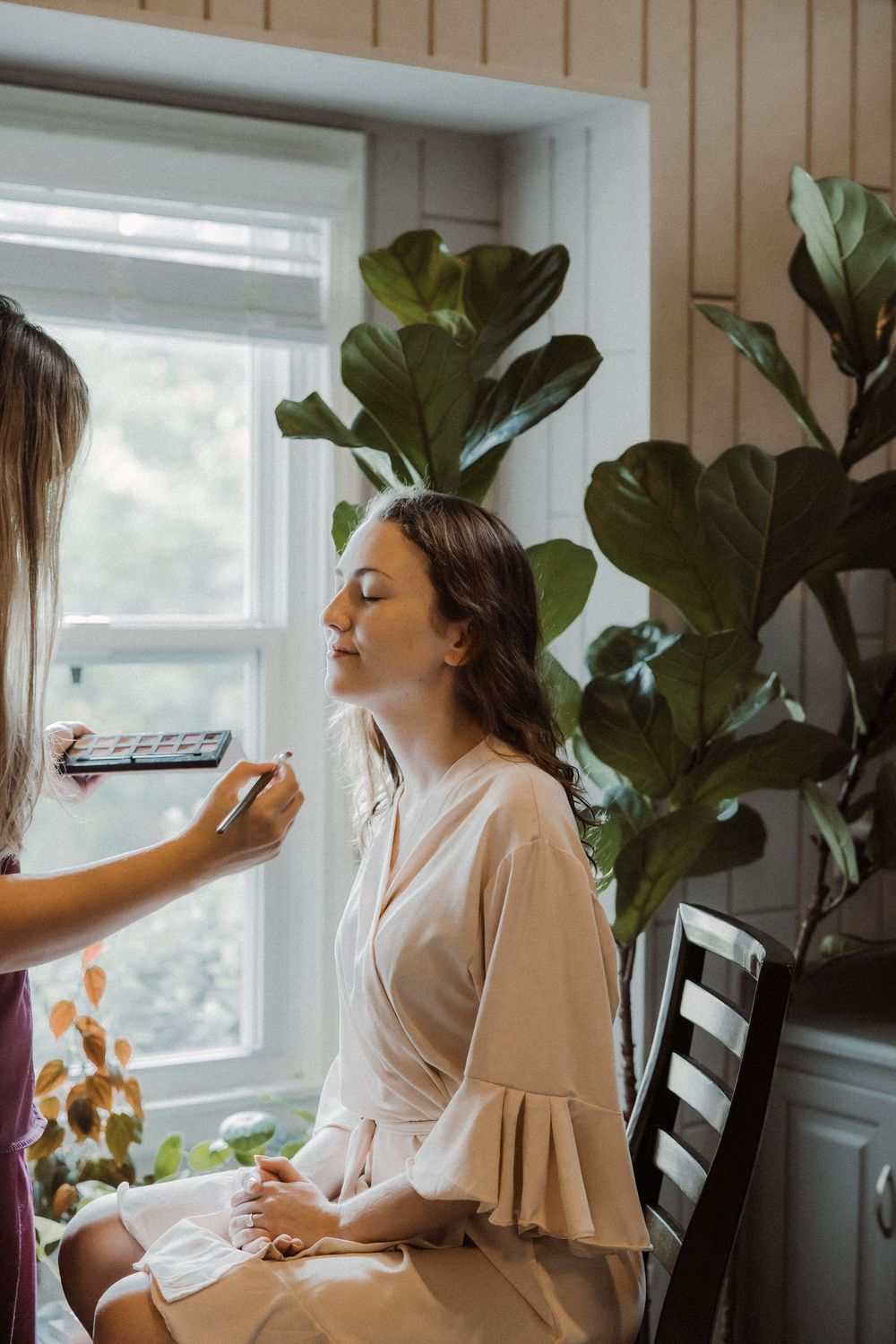 Person in white robe sits by window as makeup is applied with a large fiddle leaf fig plant in the background.