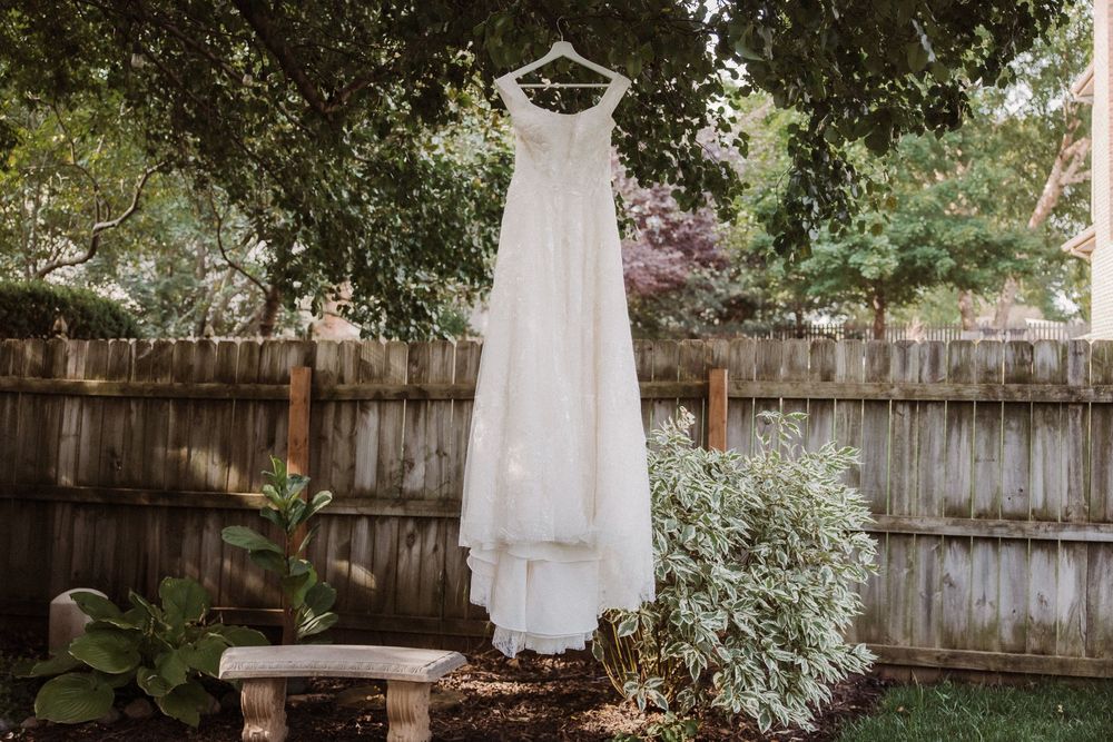 A simple white wedding dress hangs from a wooden fence in a garden surrounded by lush greenery and trees.