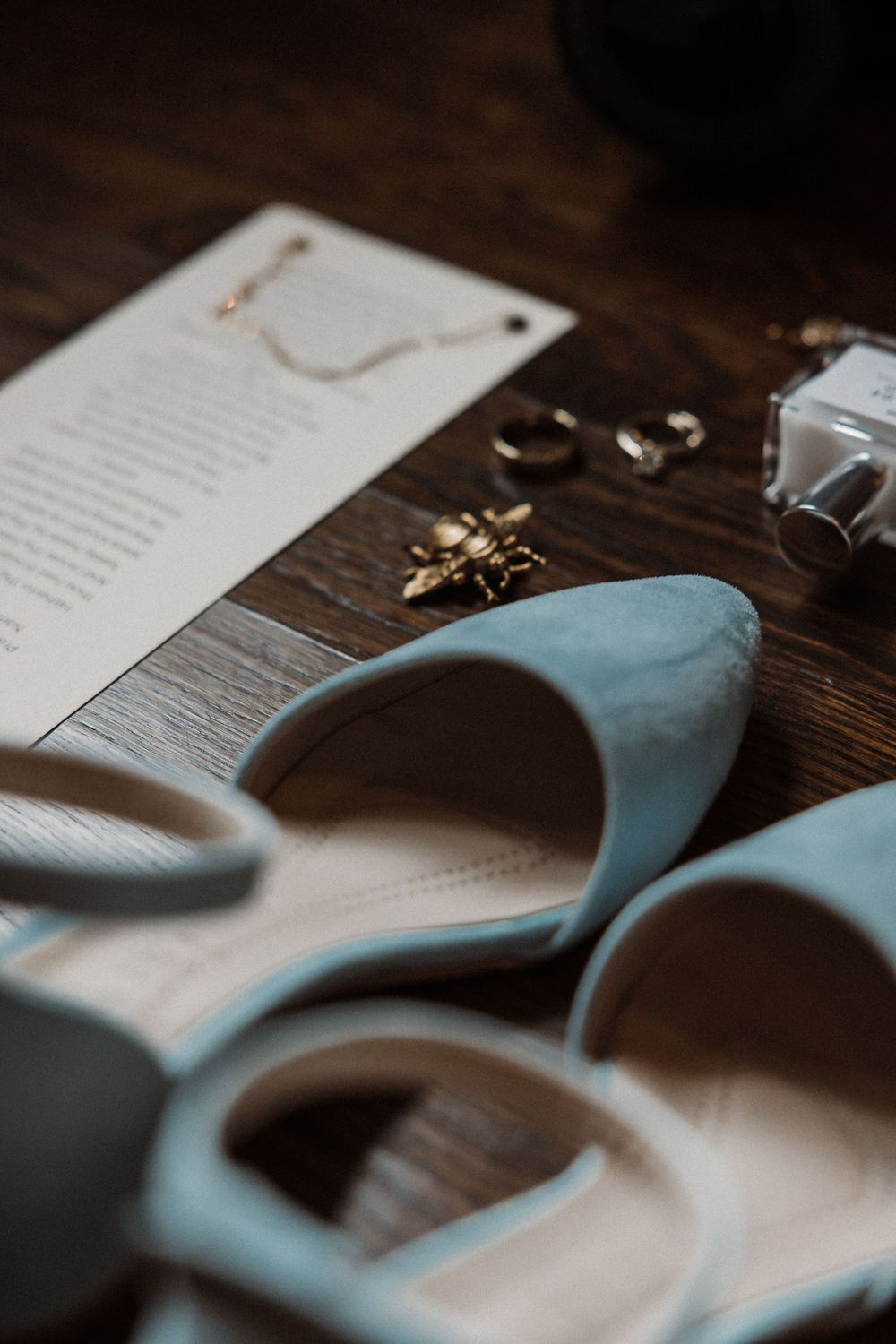 Wedding accessories including pale blue heels, rings, and invitation details arranged on a rustic wooden surface.