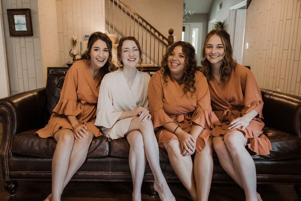 Four people wearing matching satin robes sit together on a brown leather couch while laughing and smiling.