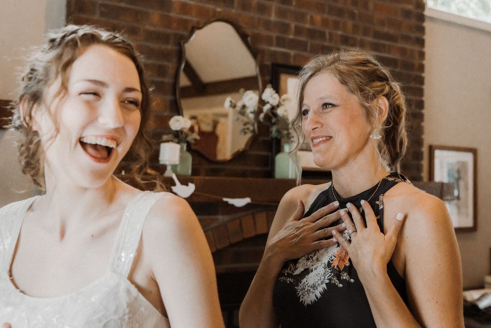 Two women laugh joyfully together during a formal event in front of a rustic brick fireplace.