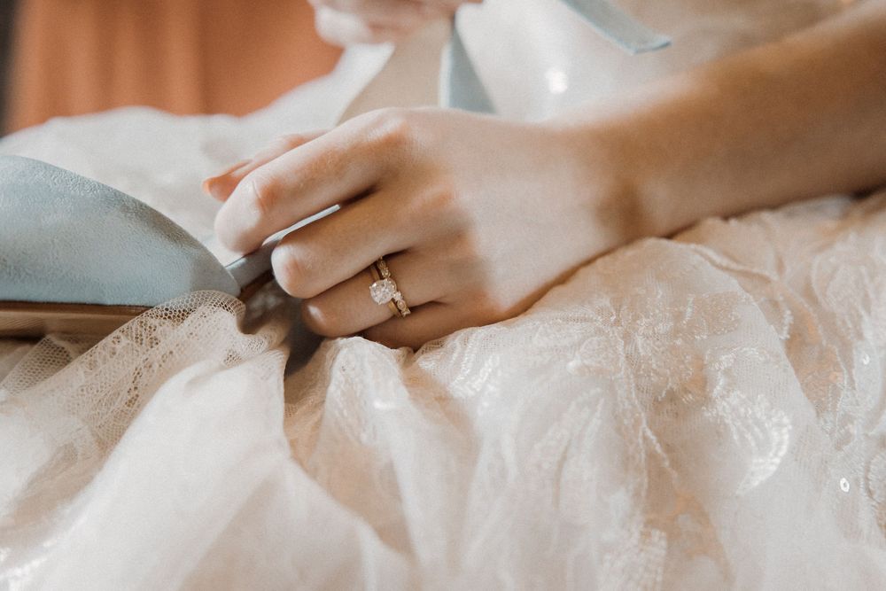 Close-up shot of elegant diamond engagement ring shown on hand resting against delicate white lace fabric.
