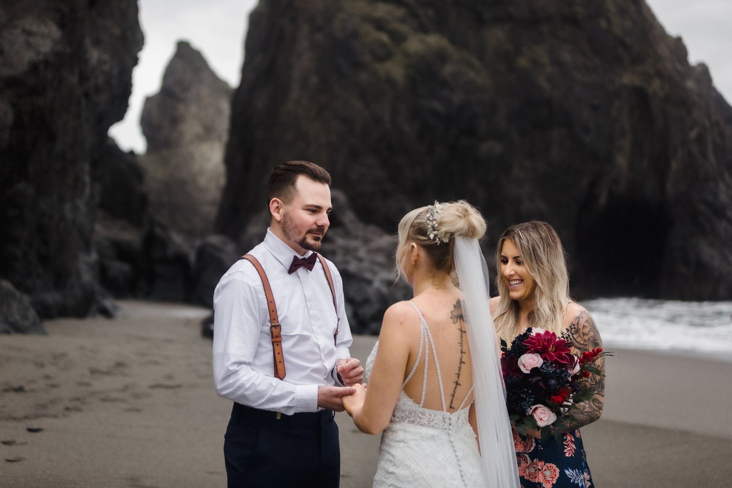 An intimate beach wedding ceremony takes place between dark rocky cliffs on a moody coastline.
