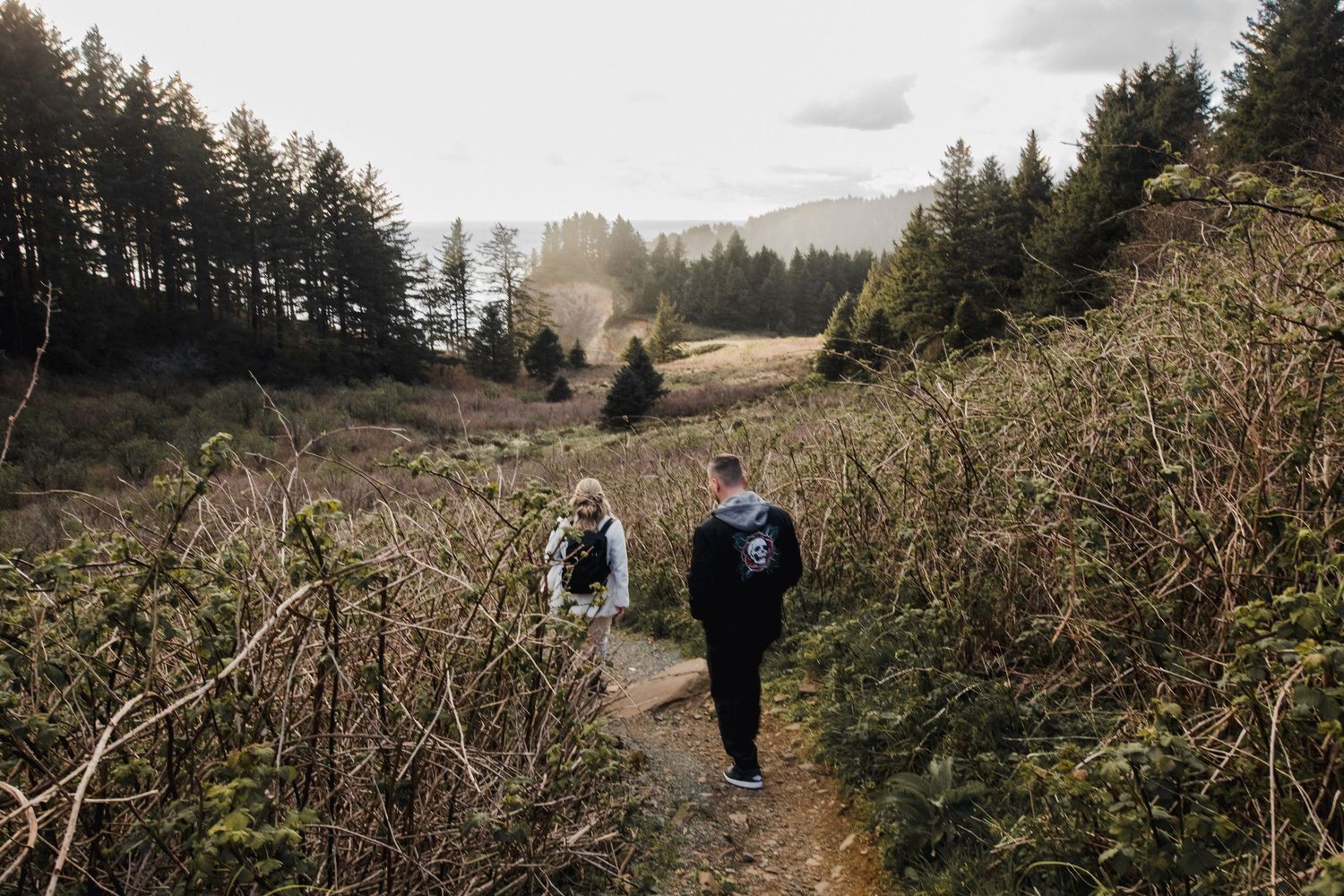 Hikers walking on a forested trail through tall grass and evergreen trees under an overcast sky.