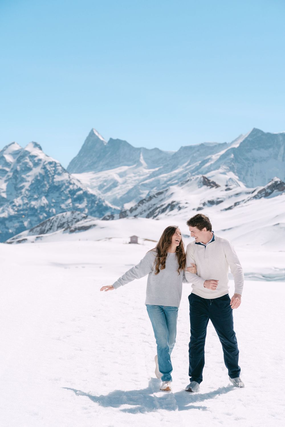 A couple walks together through deep snow with majestic snow-capped Alpine mountains rising in the background.