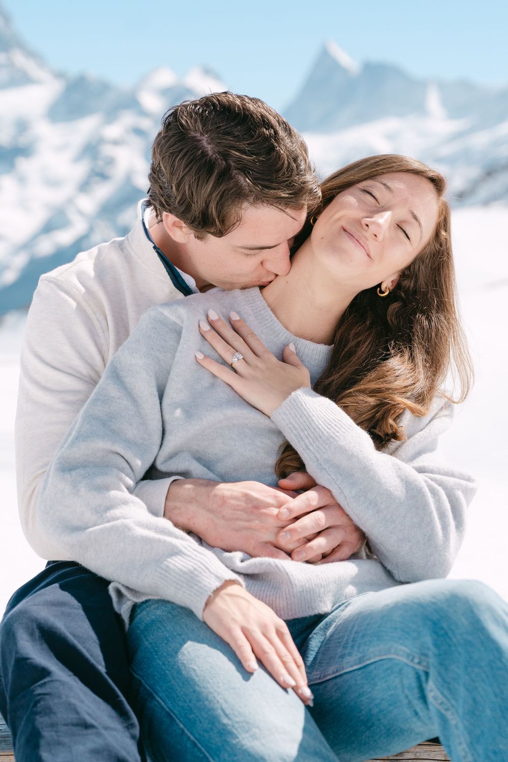 Couple sharing an intimate embrace against a snowy mountain backdrop on a bright winter day.