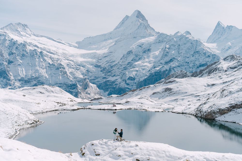 Two silhouettes stand on a snowy mountain peak overlooking a pristine alpine lake surrounded by snow-capped peaks.