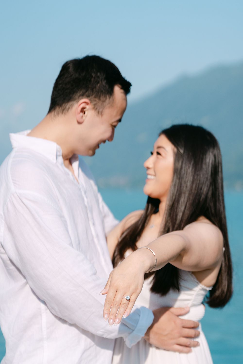 A romantic couple embraces near blue waters and mountains, sharing tender moments in a white and blue setting.
