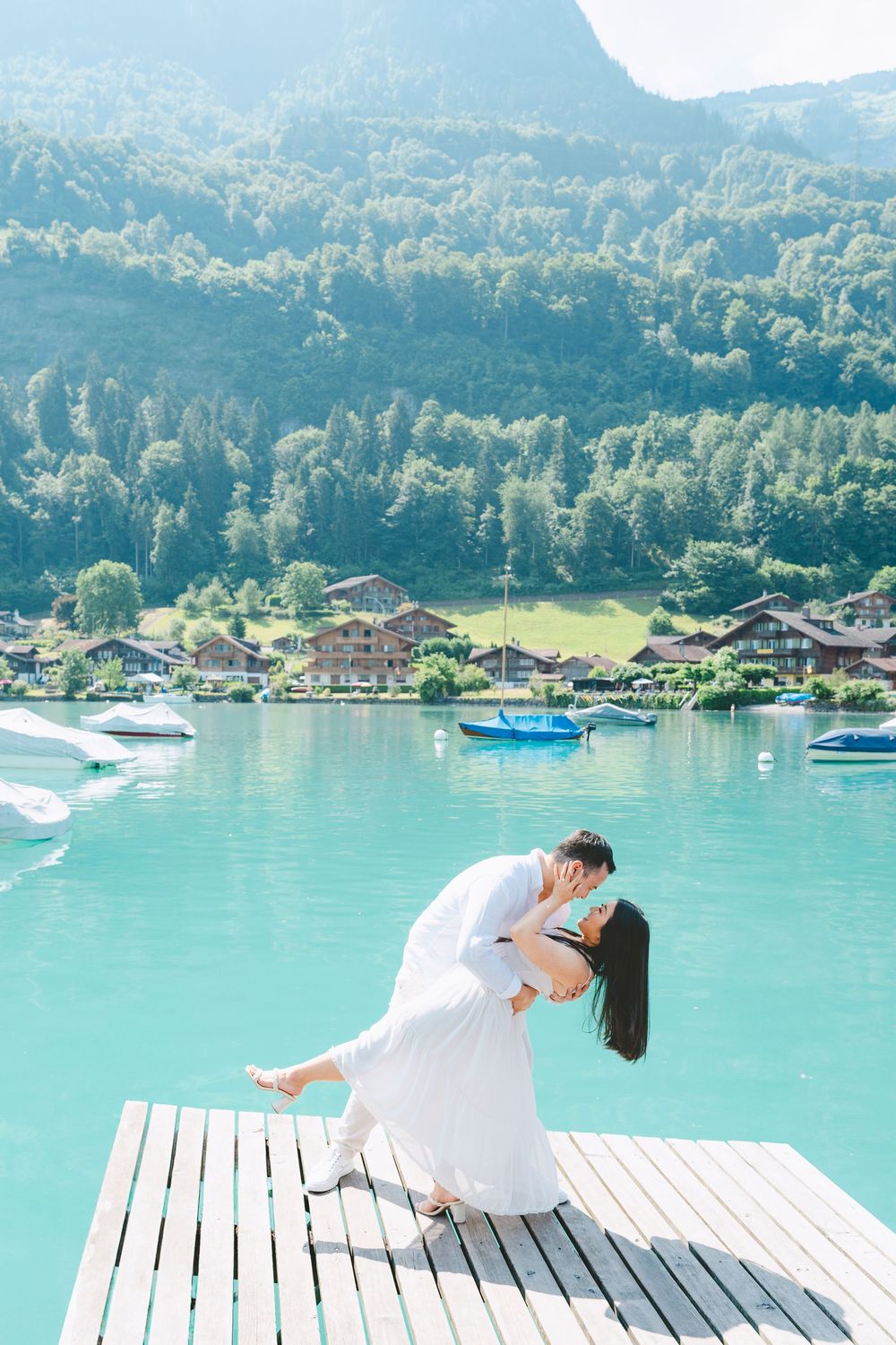 Romantic couple in white outfits poses on wooden deck overlooking turquoise lake with boats and mountains in Switzerland.