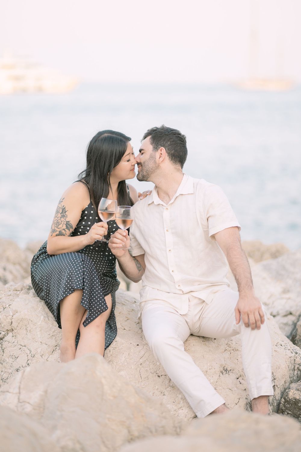 Romantic couple sharing wine while sitting on coastal rocks at sunset.