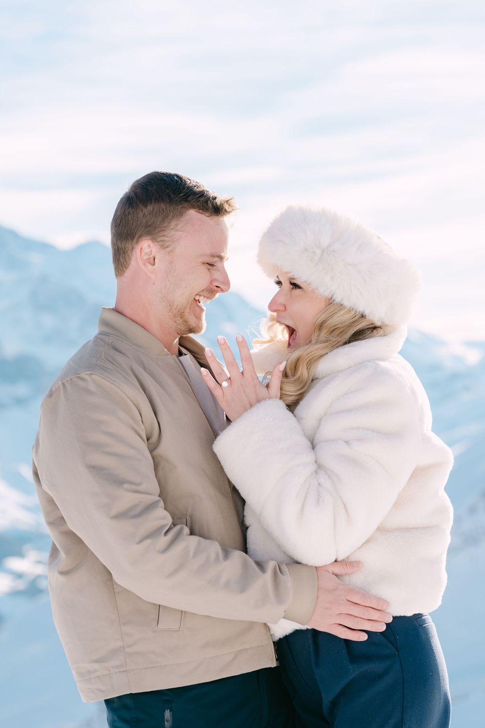 A joyful couple embraces while wearing winter coats against a snowy mountain backdrop.