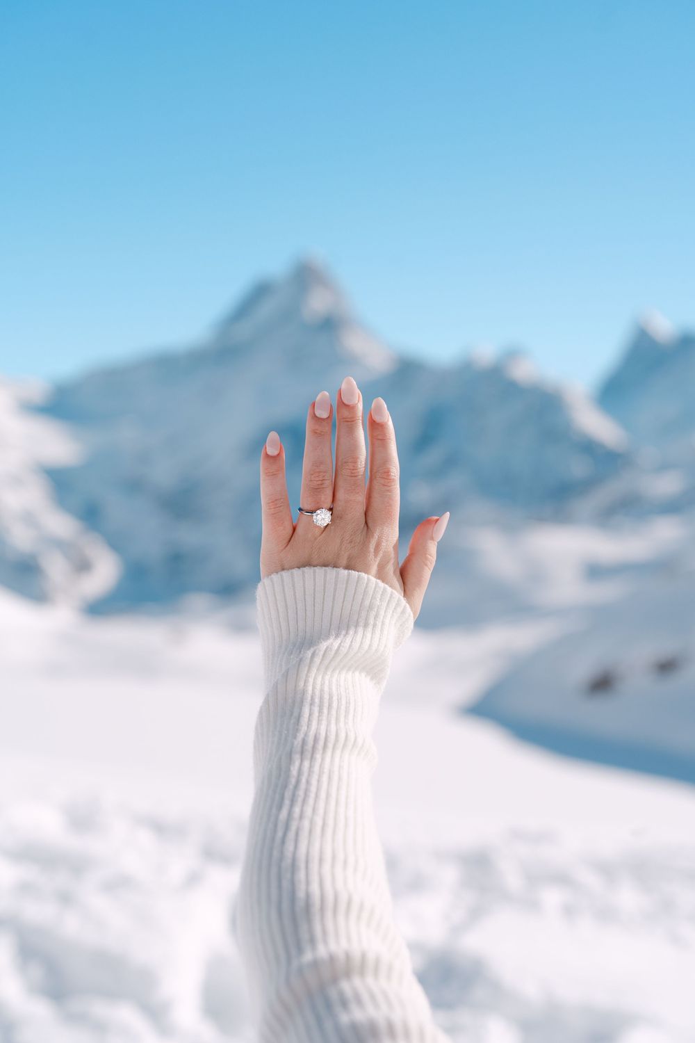 Hand wearing white sweater sleeve and engagement ring raised against snowy mountain landscape and blue sky.