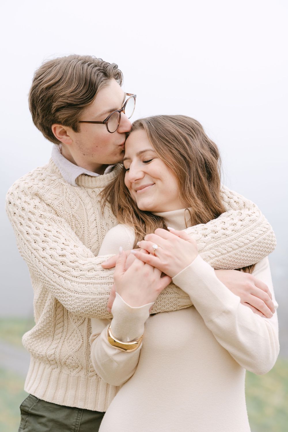 A romantic couple in cozy cream sweaters embrace tenderly outdoors on an overcast day.