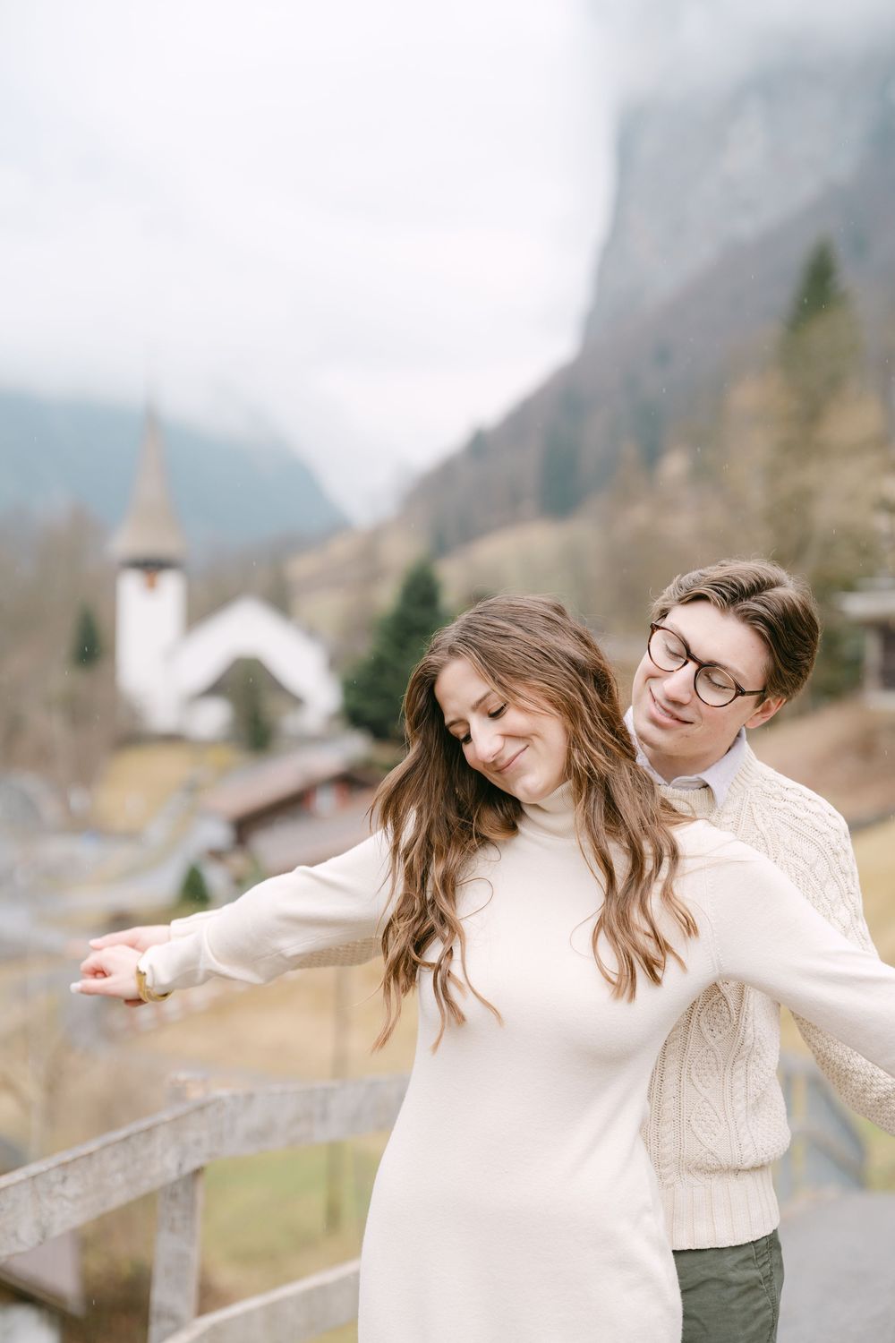 A couple in cream sweaters stand together on a mountain overlook with a quaint church steeple visible in the background.