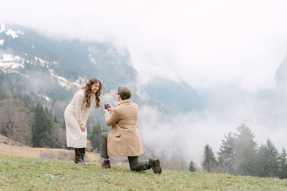 A couple shares a romantic moment on a misty mountainside meadow in a dreamy alpine landscape.