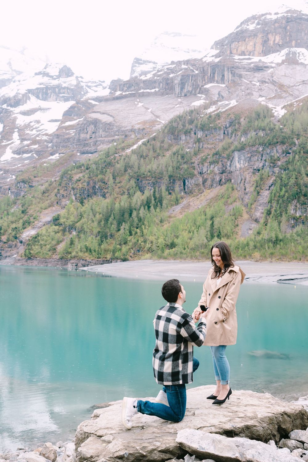 A couple sharing a romantic proposal moment on a rocky outcrop by a turquoise alpine lake with snow-capped mountains behind.
