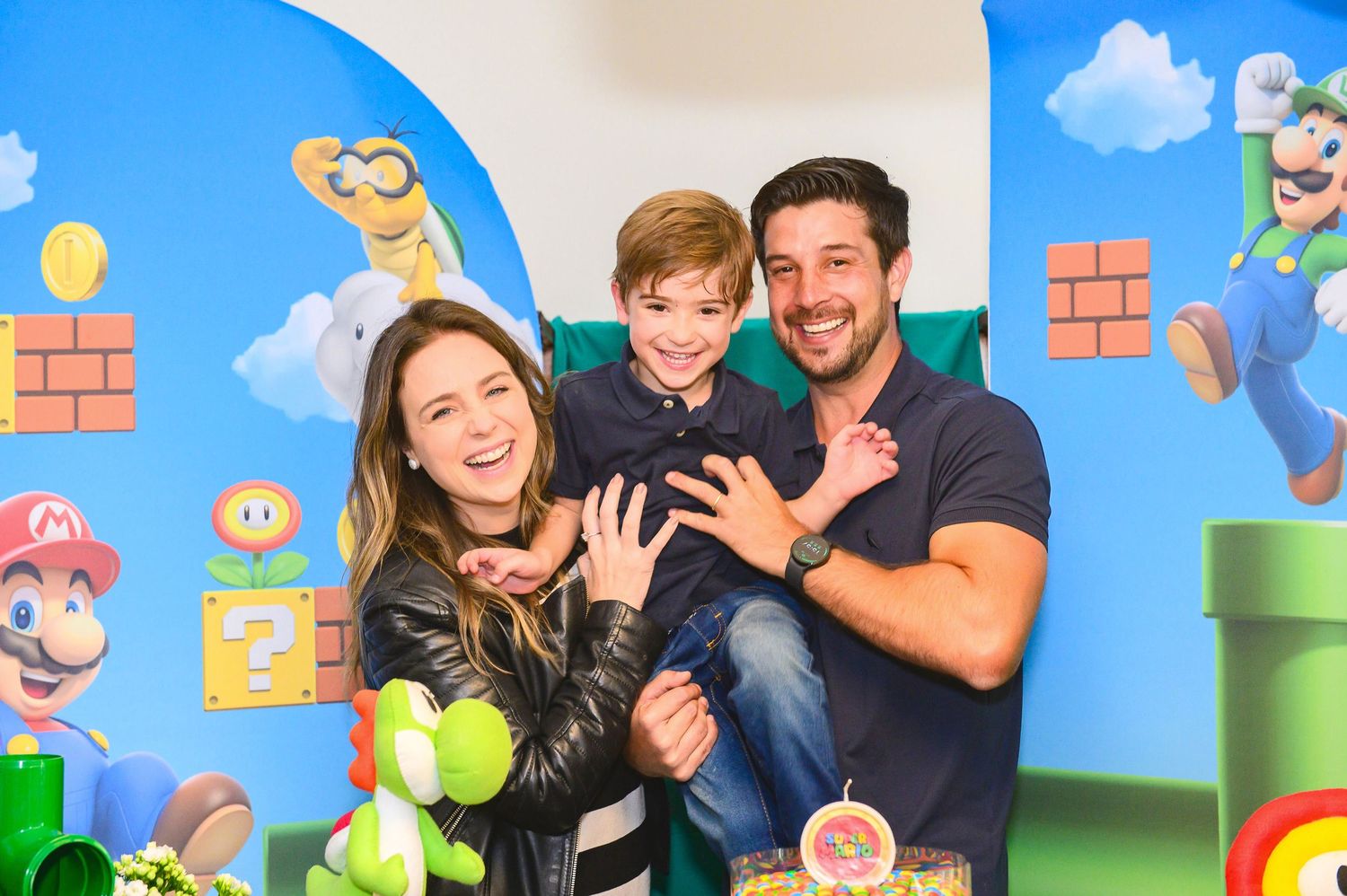A happy family poses together at a Super Mario Bros themed party with colorful video game characters on the wall behind them.