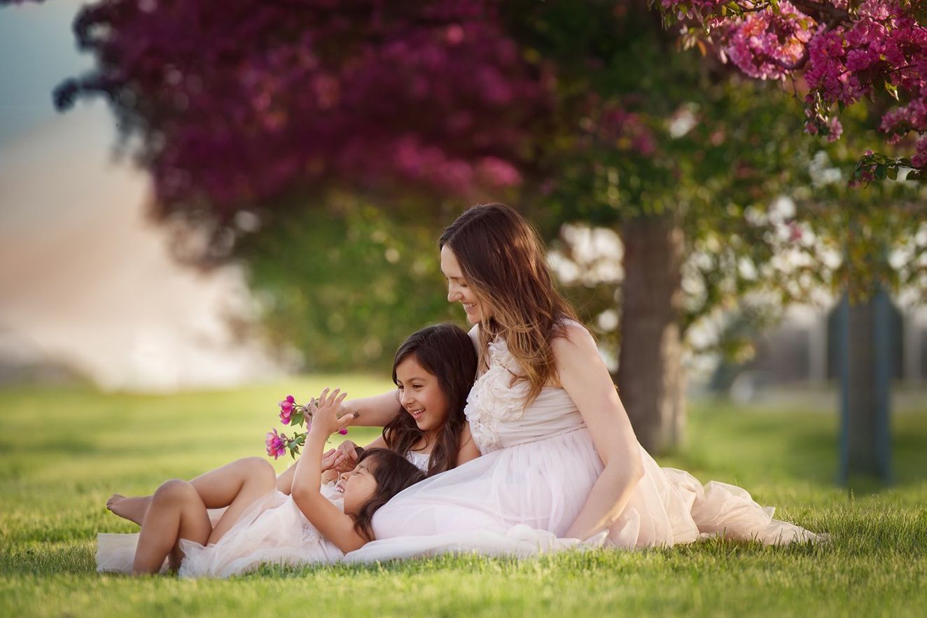 Mom with two daughters in white dresses relax under a blooming pink tree on a bright spring day in a grassy park in Palm Beach Gardens