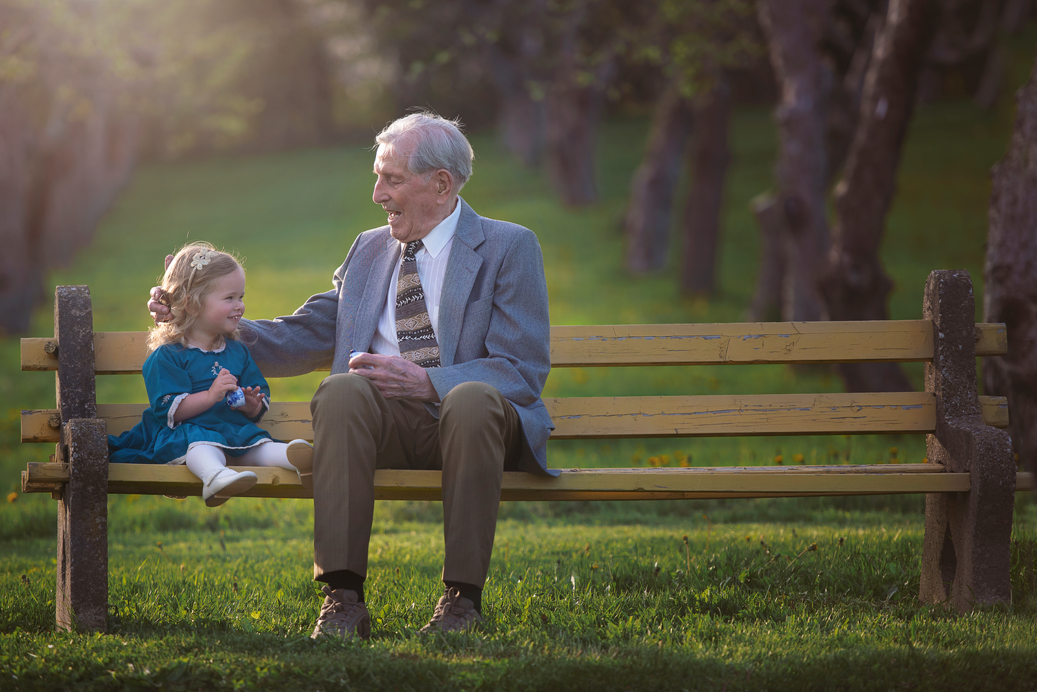 Two people sit together on a wooden park bench sharing a tender moment in golden afternoon sunlight of one of Palm Beach gardens