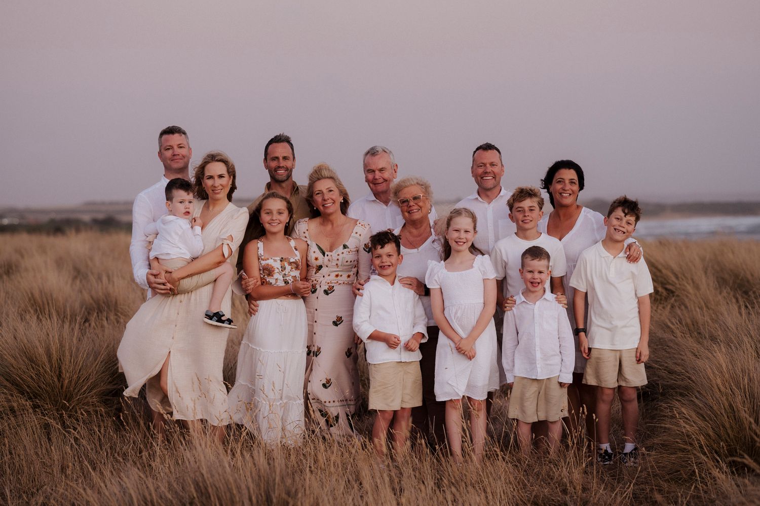 Family group in white attire posing together in tall grass against a dusky sky at sunset.