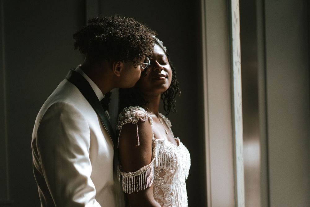 A romantic bridal portrait in natural window light shows a couple in white formal attire sharing an intimate moment.