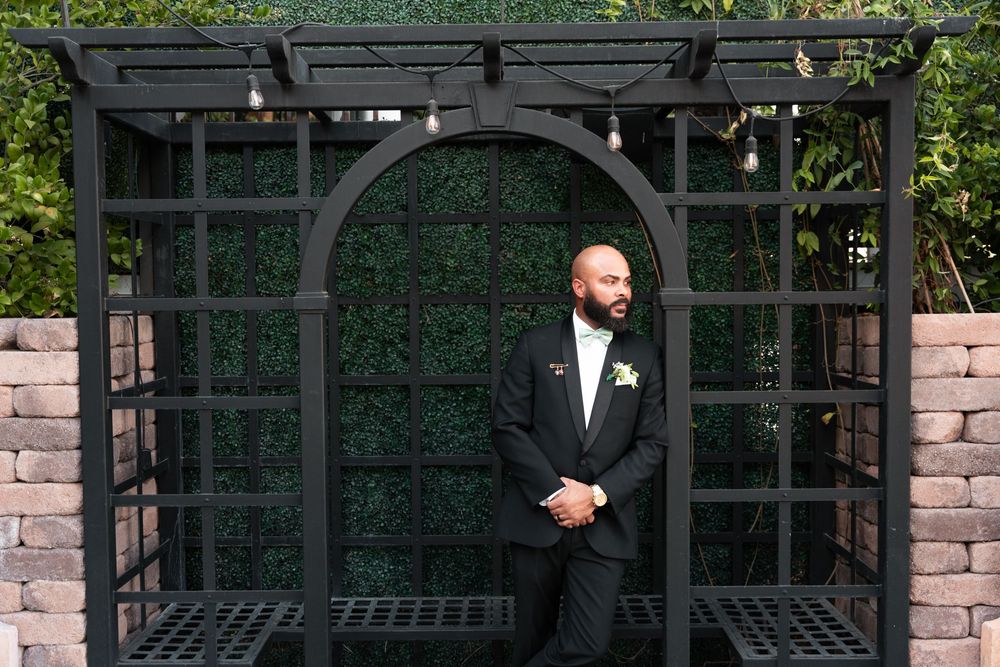 A person in a formal black suit stands in front of a dark metal garden arbor with brick walls on both sides.