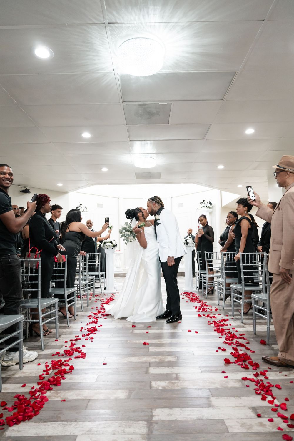 Couple sharing an intimate moment during wedding ceremony in indoor venue with red rose petals lining the aisle.
