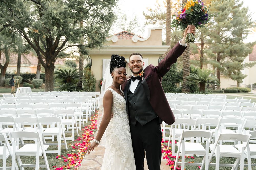 A joyful wedding couple celebrates at an outdoor ceremony venue with white chairs and rose petals lining the aisle.