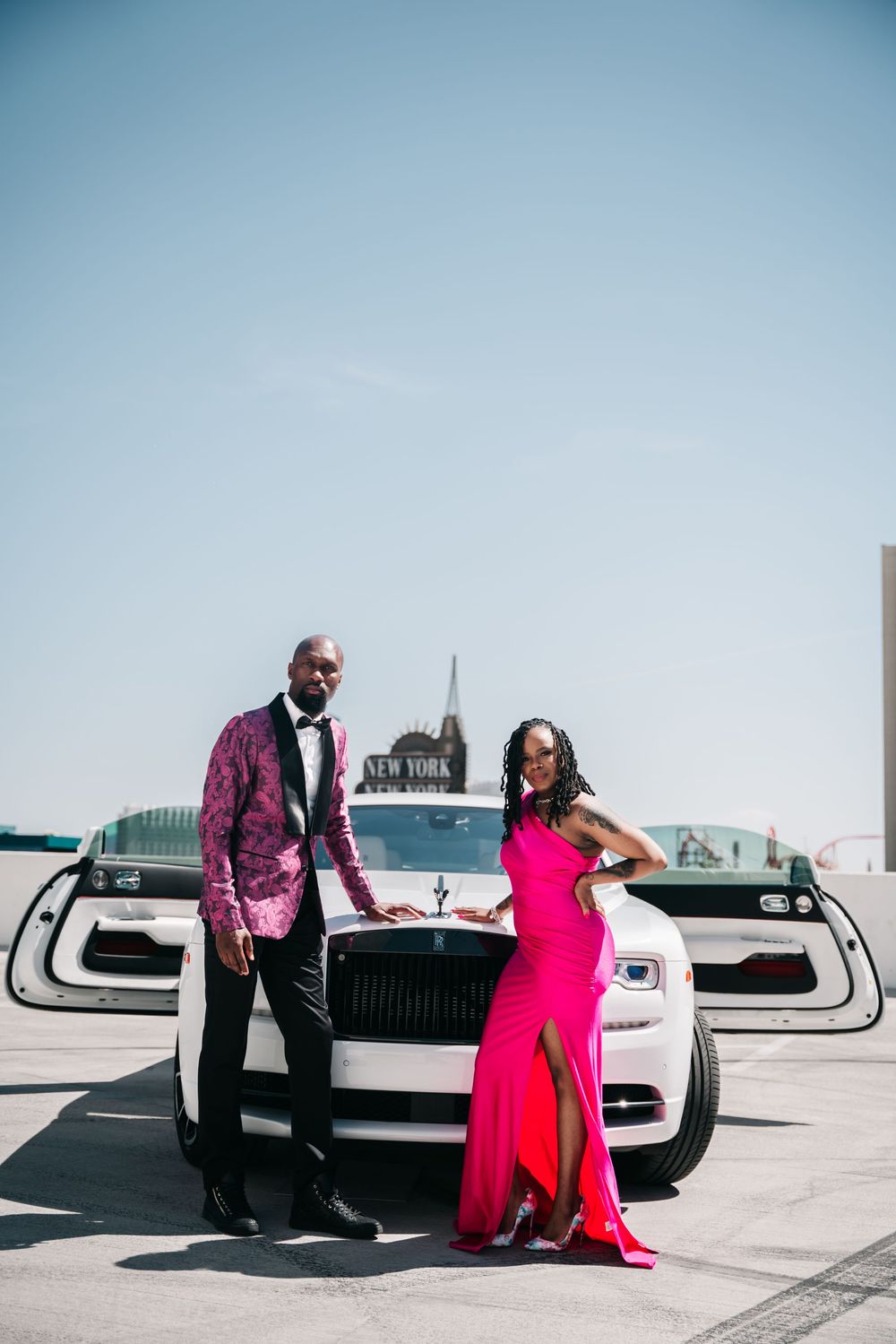 A couple poses in formal wear next to a luxurious white Rolls Royce with city skyline in background.