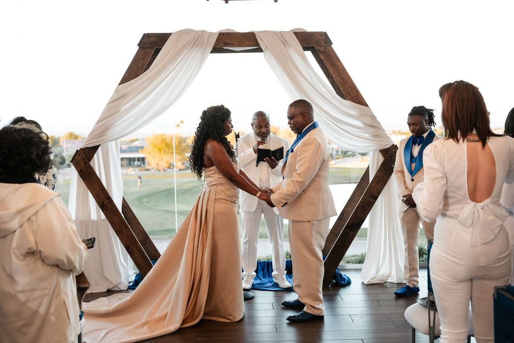 A wedding ceremony takes place under a wood hexagonal arch draped with white fabric on a covered outdoor terrace.