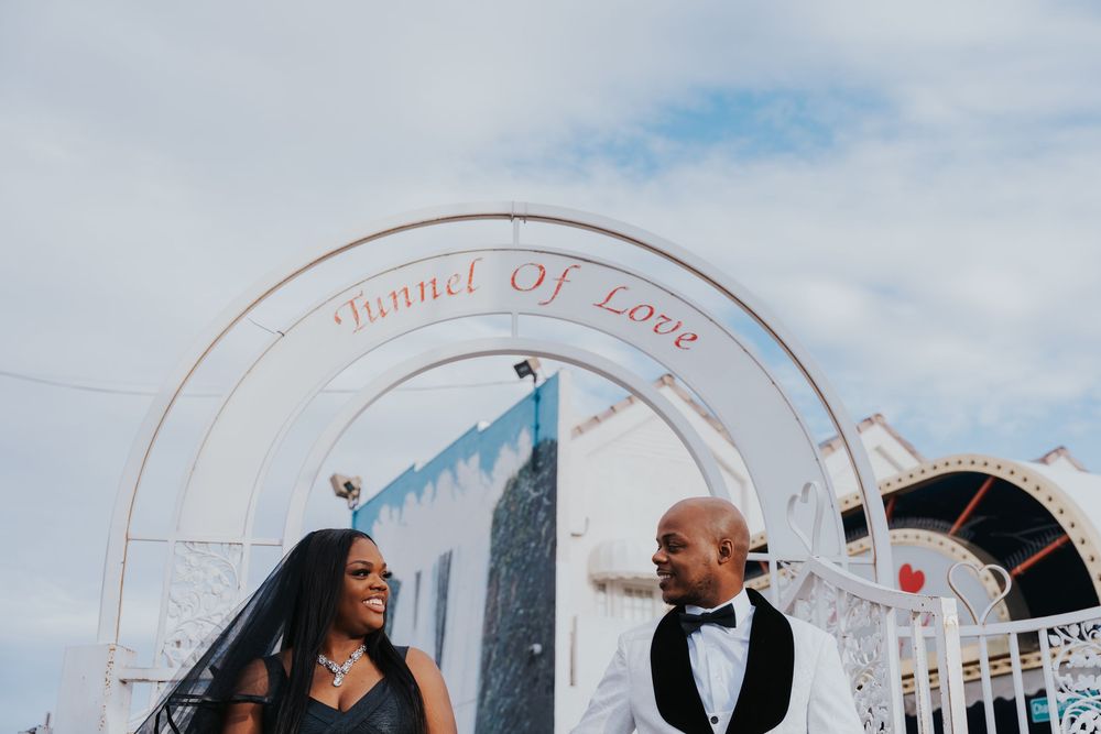 Two wedding party members laugh joyfully under a decorated outdoor arch with a blue sky backdrop.