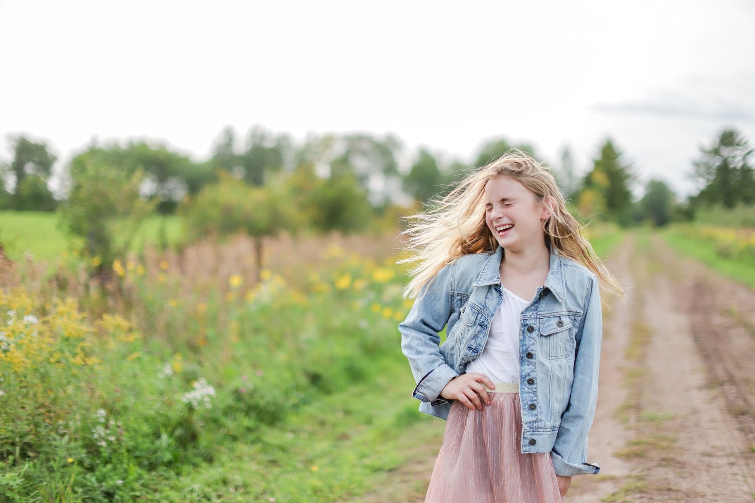 A joyful figure in a denim jacket and pink dress stands on a dirt path amid wildflowers and greenery.