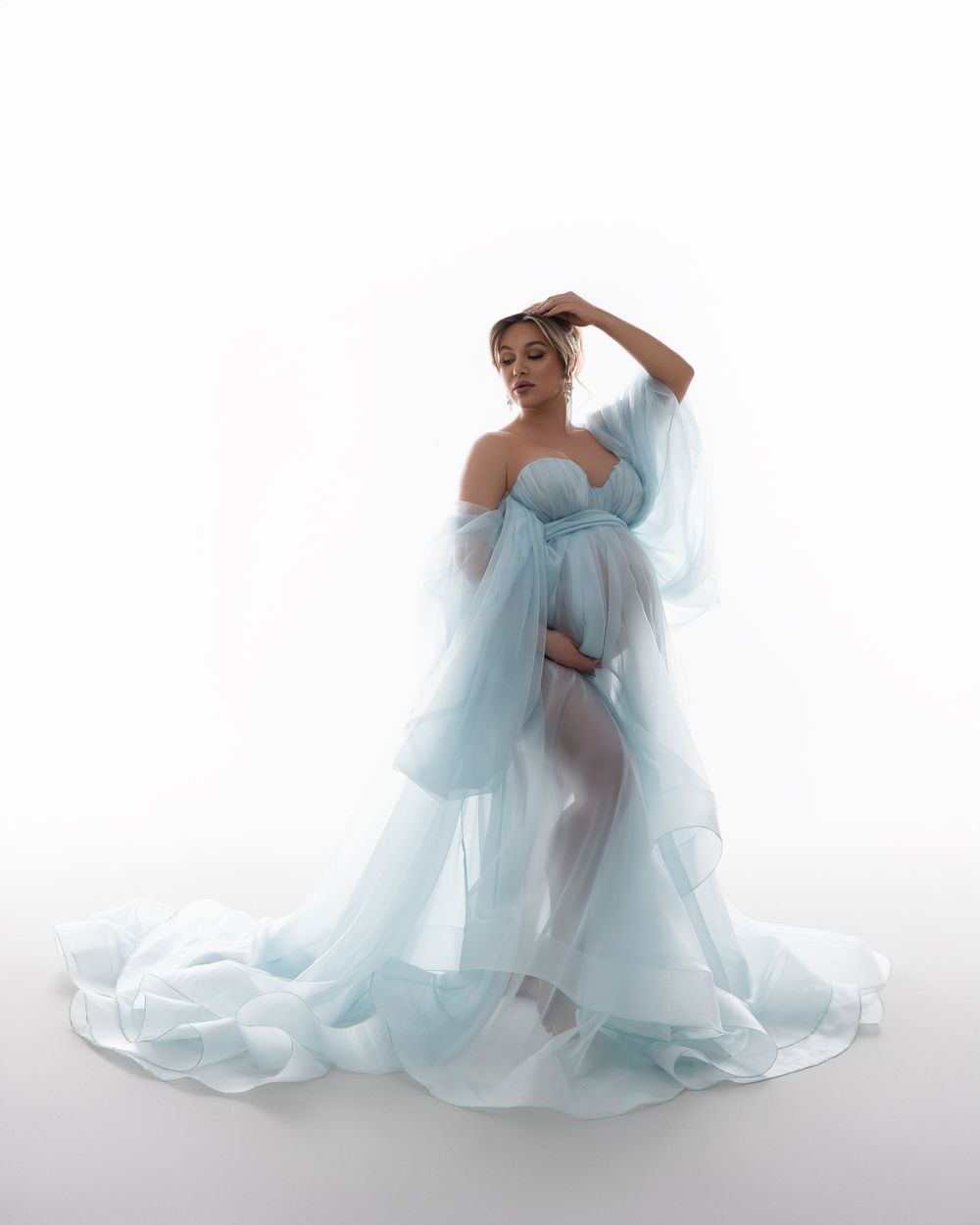 A figure in a flowing light blue tulle gown poses gracefully against a white backdrop in a dreamy fashion portrait series.