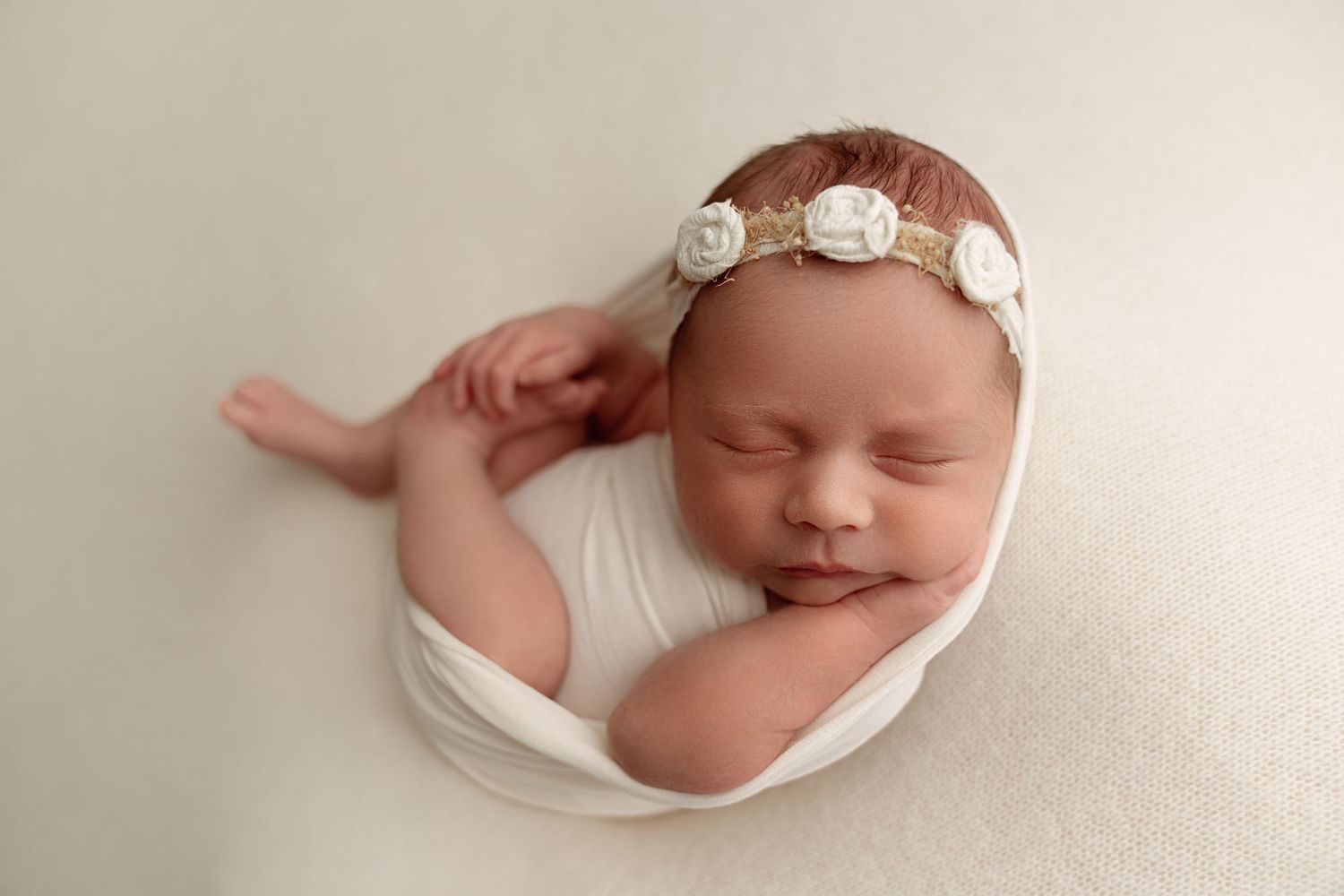 A peaceful sleeping newborn swaddled in cream fabric lies in a round bowl on rustic wooden planks with white flowers nearby.