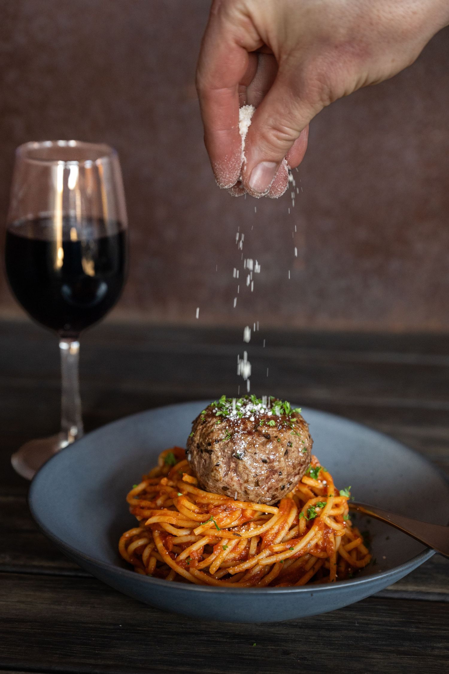 Hand sprinkles seasoning over a meatball served atop spaghetti in tomato sauce with red wine glass in background.