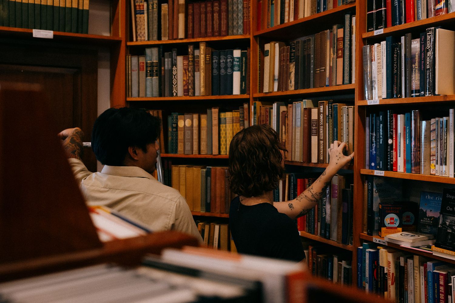 Two people browse books on wooden shelves in a cozy library setting with warm lighting.