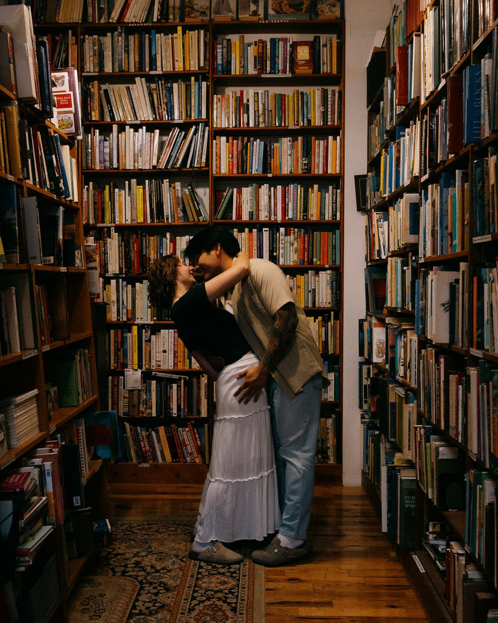 A couple dramatically kissing at the end of an aisle in a bookstore.