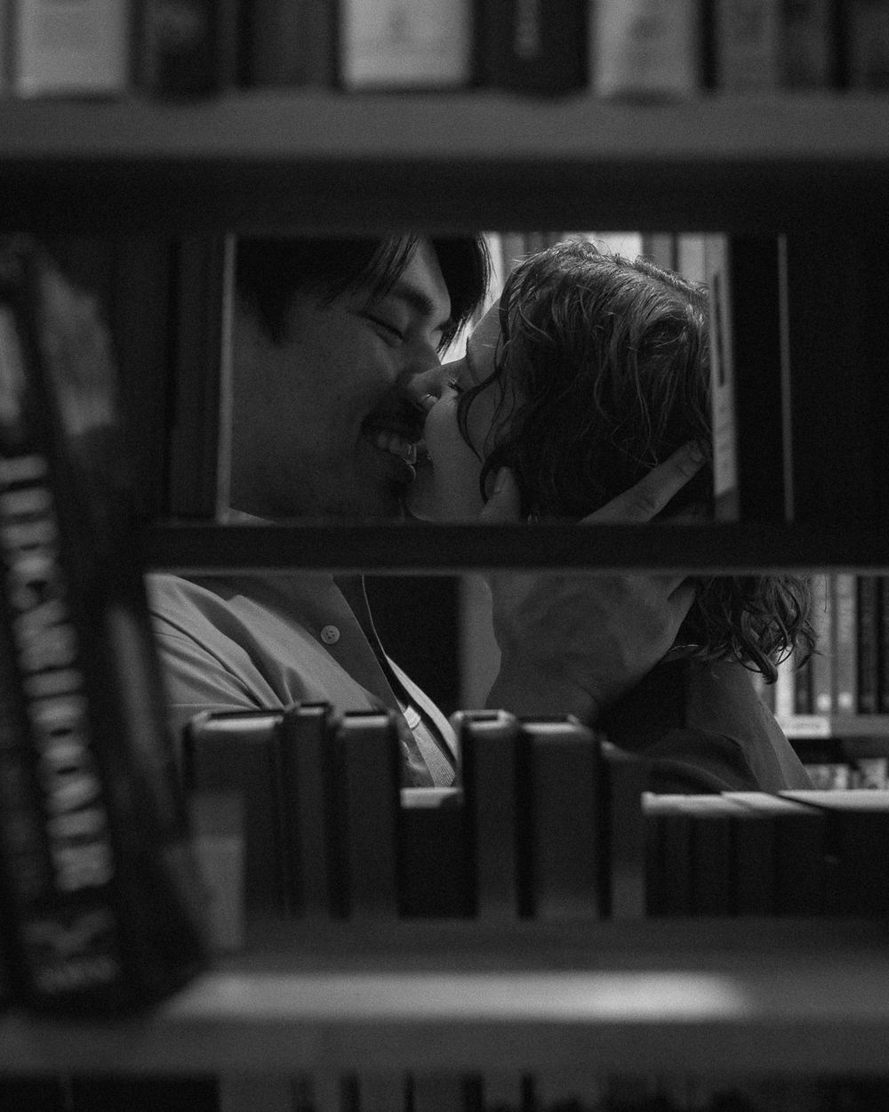 A black and white photo of a couple kissing on the other side of a bookshelf, as shot through the shelf.