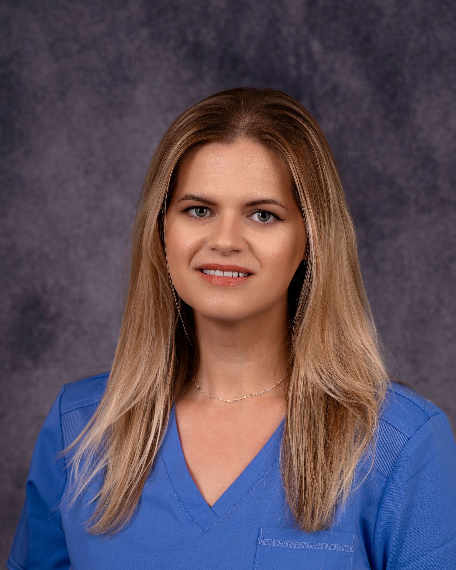 Professional medical worker in blue scrubs poses against grey studio backdrop.