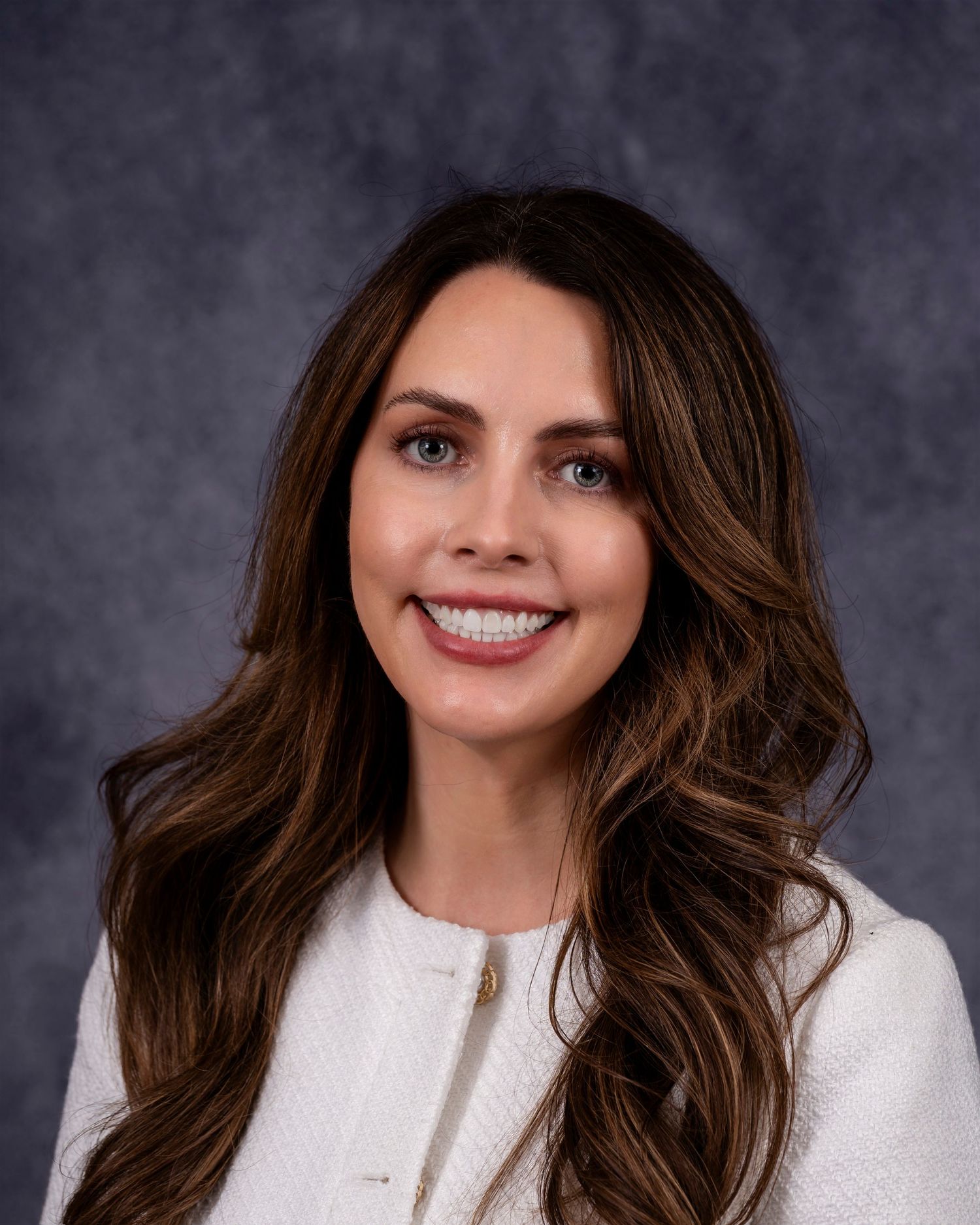 Professional headshot of someone with warm brown wavy hair wearing a white blazer against a gray background.