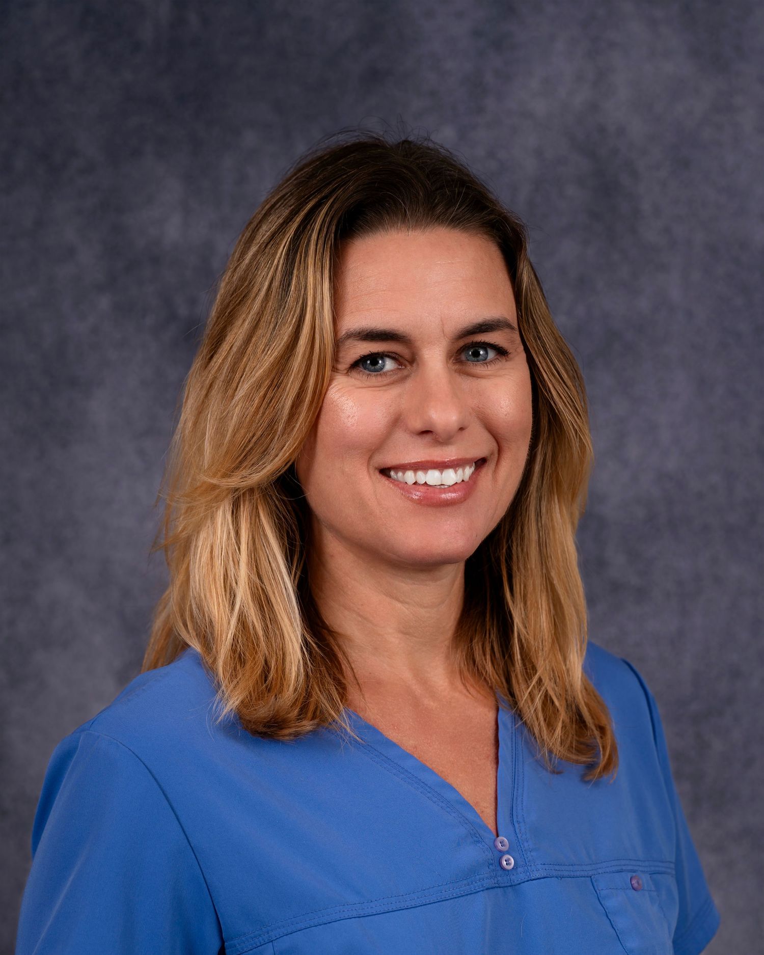 Professional portrait of a healthcare worker smiling against a gray background wearing a blue medical scrub top.