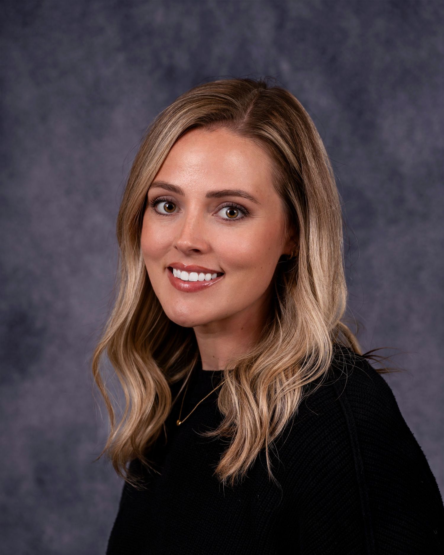 Professional headshot portrait of a person with blonde wavy hair wearing a black top against a gray background.