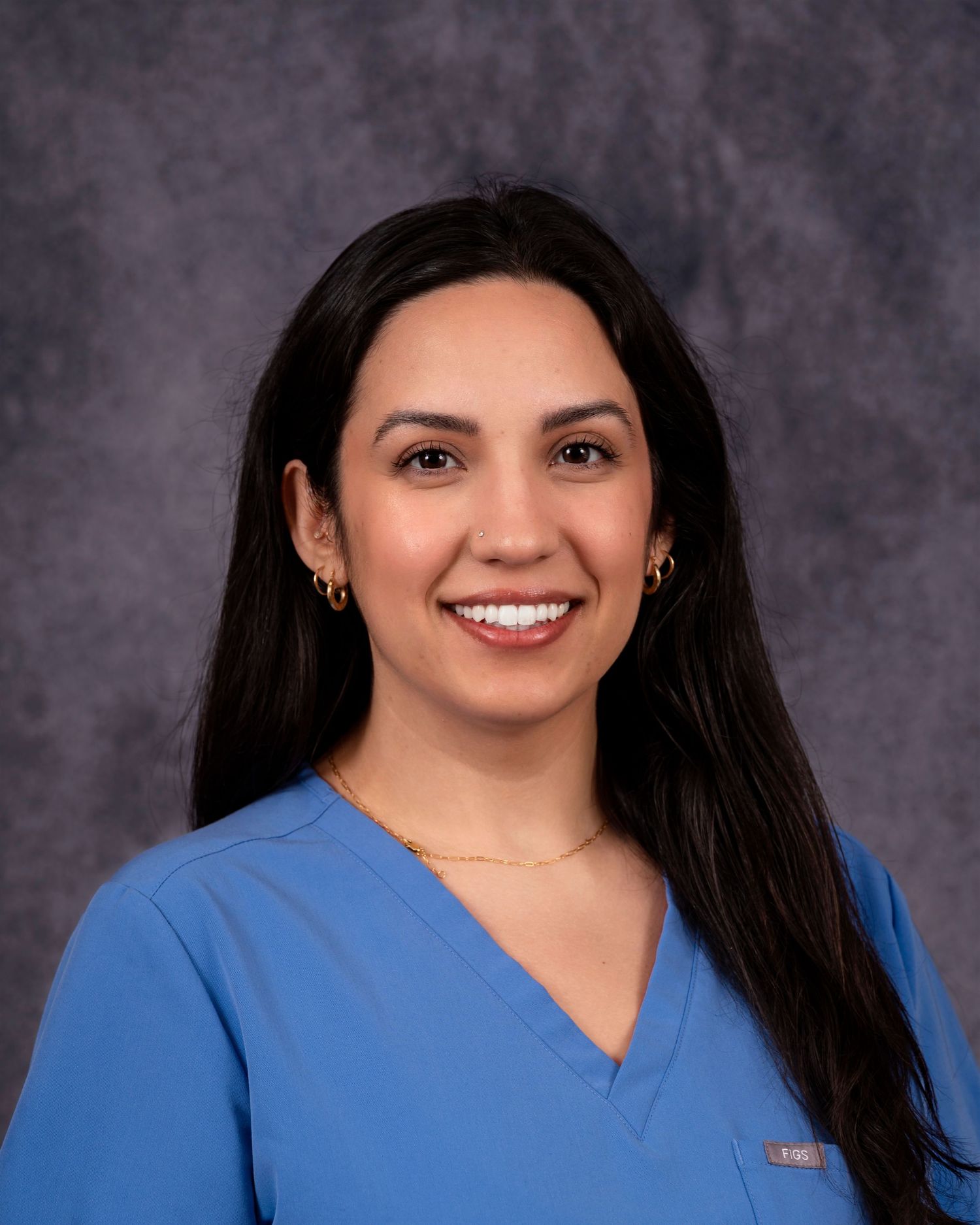 Professional headshot of a healthcare professional with long dark hair wearing blue medical scrubs.